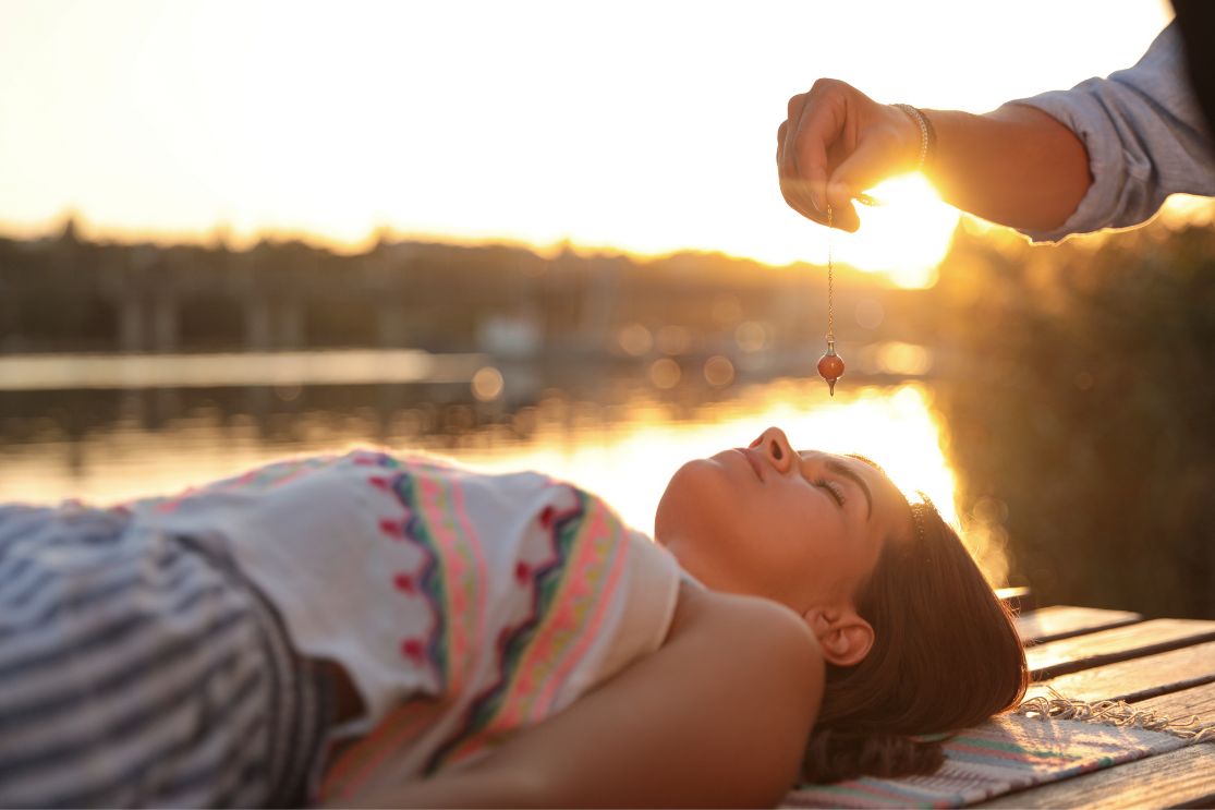 Tranquil energy retreat setting by the river, with a woman lying on a yoga mat and another woman holding a dosa above her forehead